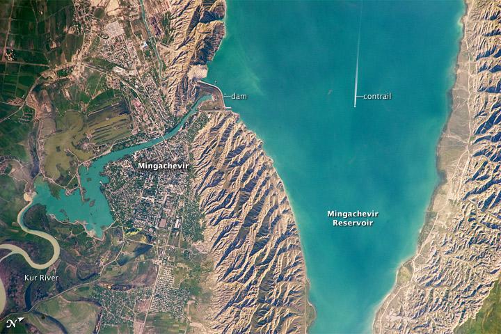 This astronaut photograph highlights the southern Mingachevir Reservoir in north-central Azerbaijan. The Mingachevir Reservoir occupies part of the Kura Basin, a topographic depression located between the Greater Caucasus Mountains to the northeast and the Lesser Caucasus Mountains to the southwest. The afternoon sun highlights distinctive parallel patterns in the hills that are the result of water and wind erosion of different rock layers exposed at the surface. The nearby city of Mingachevir (left) is split by the Kur River after it passes through the dam and hydroelectric power station complex at image top center. The current city was built in support of the hydroelectric power station constructed as part of the then-Soviet Union's energy infrastructure for the region. Today, Mingachevir is the fourth-largest city in Azerbaijan (by population), and it has become a cultural and economic center of the country. The width of the reservoir illustrated here is approximately 8 km (5 mi); a jet flying over the reservoir left a contrail midway between the shorelines. Photo courtesy of NASA.