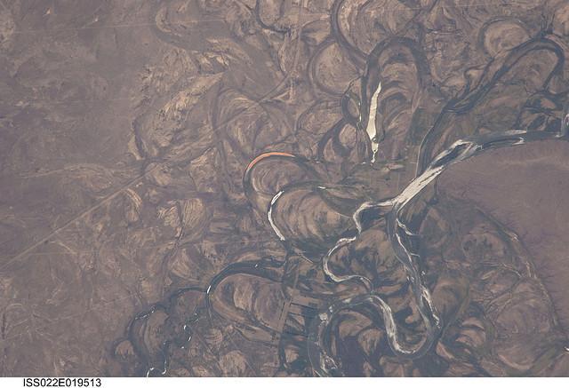 The Rio Negro in Patagonia is recognizable from space as one of the most meandering rivers in South America. This phenomenon is well illustrated in this view, where the entire river floodplain (approximately 10 km (6 mi) wide) is covered with curved relics of channels known as meander scars. The main channel of the river, flowing south at this point 60 km (36 mi) south of the city of Choele Choel (not shown) appears in partial sun glint at right. Sun glint occurs when light is reflected off a water surface directly back towards the viewer, imparting a silvery sheen to those areas. When meander scars contain water they are known as oxbow lakes, some of which are also highlighted by sun glint in the image. Meander scars show the numerous past positions of river bends, produced as the river snaked across the plain in the very recent geological past. The Rio Negro is a dramatic example of how mobile a river can be. The orange tint to the water in one of the oxbow lakes (center) could result from orange salt-loving algae. Image courtesy of NASA.