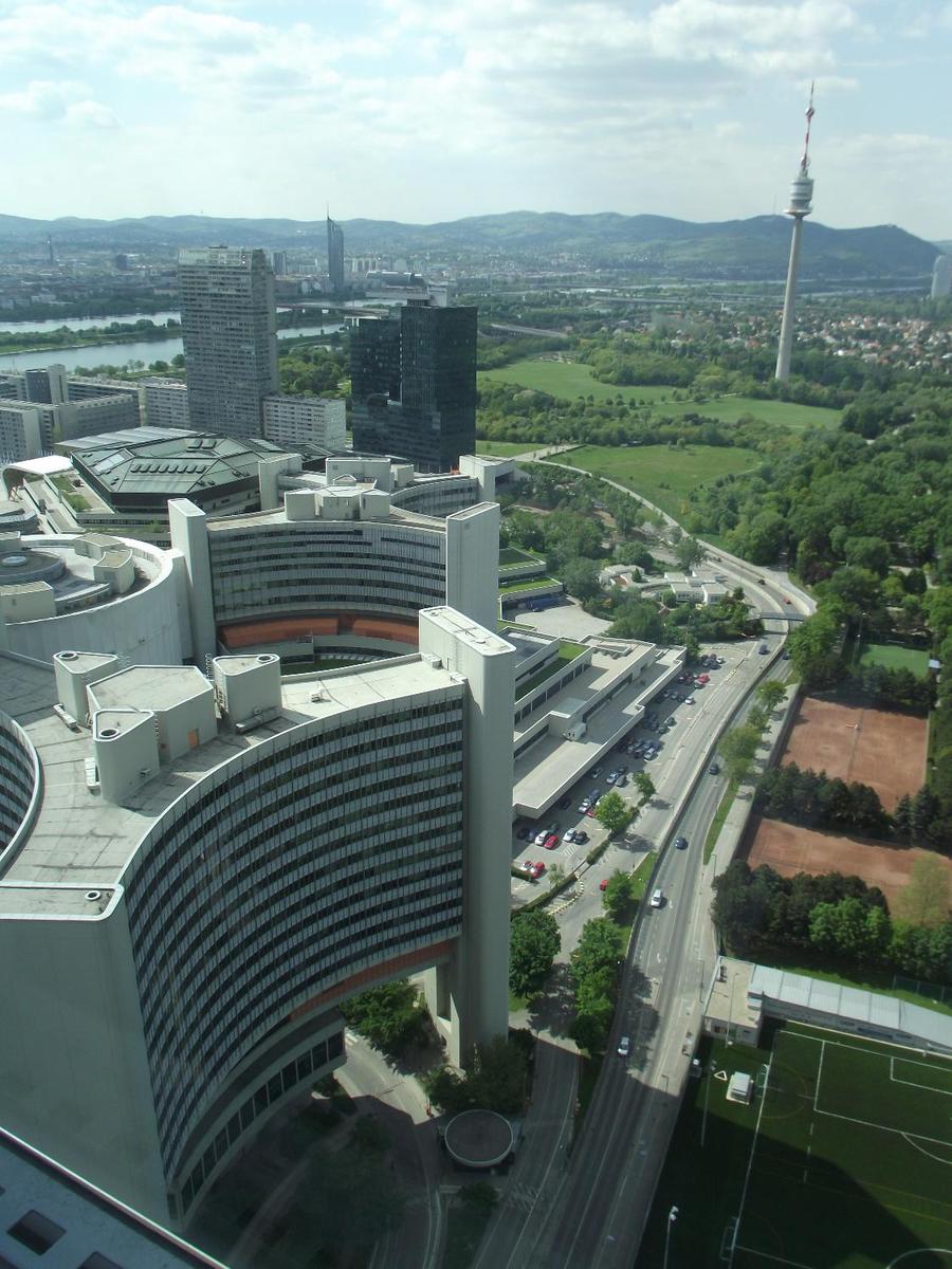 A bird's-eye view of two of the six Y-shaped office buildings, as well as the central cylindrical conference center, that make up part the United Nations Office complex in Vienna. In the distance is the 252 m (827 ft)Donauturn (Danube Tower) the tallest structure in Austria. In addition to its use as a communication tower, it also houses two revolving retaurants and has an observation deck.