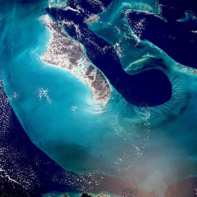 An even more stark view of the dark blue Tongue of the Ocean whose depths reach up to 1,800 m (6,000 ft). By comparison, the waters of the Bahama Platform (light blue) are frequently less than 15 m (50 ft) deep. They are warm and become extremely salty due to evaporation and limited circulation from the open ocean. Crystals of aragonite, a calcium carbonate mineral derived from the shells of single-celled marine organisms, and direct precipitation, form into oolites (small spherical grains of limestone) as the tidal currents swirl back and forth. Lithification of the carbonate sands produces an oolite limestone. Although the water is warm and clear, corals do not live in these shallows, probably because of the salt content. Though chemically very similar, the rocks resulting from this precipitating process have a quite different origin from those formed from coral reefs. Image courtesy of NASA.