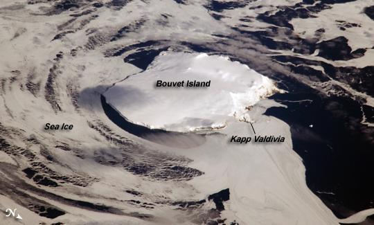 Bouvet Island is one of the most remote islands in the world; Antarctica, over 1,600 km (1,000 mi) to the south, is the nearest land mass. Located near the junction of three tectonic plates, the island is mostly formed from a shield volcano that is almost entirely covered by glaciers. The prominent Kapp (Cape) Valdivia on the northern coastline is a peninsula formed by a lava dome. It is only along the steep cliffs of the coastline that the underlaying dark volcanic rock is visible against the white snow and ice blanketing the island. Photo courtesy of NASA.