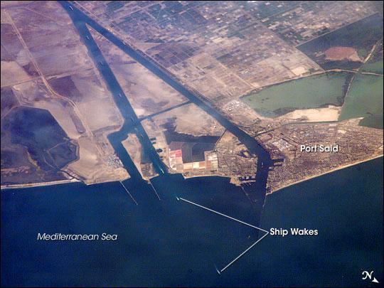 This oblique image captures a northbound convoy of cargo ships entering the Mediterranean Sea from the Suez Canal (leftmost canal branch at image center). The Canal connects Port Said on the Mediterranean Sea with the port of Suez on the Red Sea, and provides an essentially direct route for transport of goods between Europe and Asia. The Canal is 163 km (101 mi) long and 300 m (984 ft) wide at its narrowest point - sufficiently wide for ships as large as aircraft carriers to traverse it. Transit time from end to end averages 14 hours. Image courtesy of NASA.