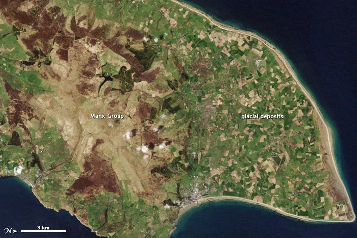 The Isle of Man is a small island with a complex geologic history. Geologists have studied and reported on the Isle of Man since the 19th century, making it a popular destination for rock hounds. Roughly 50 km (30 mi) from northeast to southwest, the island preserves rock layers dating back hundreds of millions of years. While most of the rocks are covered by soil, some rock layers exposed at the surface, along the coast, and in quarries have shed light on this island's assorted ancient landscapes.
This natural-color satellite image of the Isle from 1 May 2001 shows the northern end of the island; the image has been rotated so north is to the right. Croplands cover the relatively flat terrain of the northern coastal plain, which forms a rough triangle. Underlying the fields are glacial sediments. Between 70,000 and 10,000 years ago, a giant ice sheet covered the Isle of Man. The ice advanced and retreated multiple times, occasionally piling up rocks to form hills. As the ice melted, all the dirt and debris locked within it came to rest on the northern plain.
Toward the south, the land rises. The rock layers in this region are collectively known as the Manx Group; they make up the bulk of the Isle of Man, and comprise a mixture of sedimentary and volcanic rocks, folded and faulted by millions of years of tectonic pressures. They are far older than the glacial sediments coating the northern plain, having been formed between 490 million and 470 million years ago at the bottom of an ancient sea floor. Visible from the sky, the uneven contours of these rock layers hint at their complicated history. Photo courtesy of NASA.