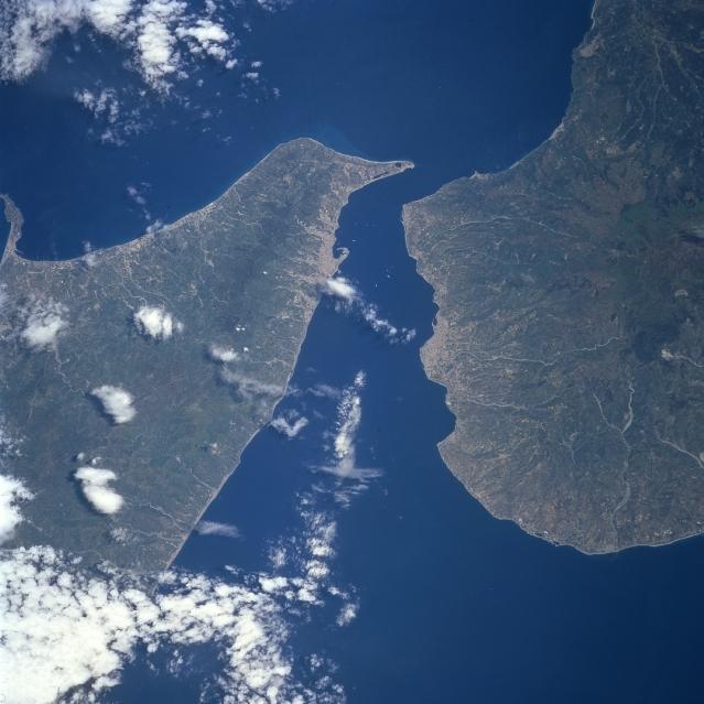 The Strait of Messina separates Sicily (left-west) from the mainland of Italy (right-east) and also provides a water passage, connecting the Tyrrhenian Sea with the Ionian Sea. The landscape on both sides of the strait is characterized by rugged mountainous terrain, short-flowing rivers, and clustered urban areas along the narrow coastline. The concentration of lighter colored areas along the shoreline identifies the location of Messina (Sicily) and Reggio di Calabria (Italy). Image courtesy of NASA.