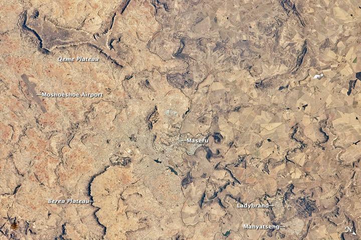 Maseru is the capital city of the Kingdom of Lesotho, and is located along the northwestern border of the country with the Republic of South Africa. The footprint of the urban area, recognizable by street grids and distinctive blue-roofed industrial buildings at image center, is only just visible against the surrounding landscape. The city has expanded eightfold since independence from the United Kingdom in 1966, and is now home to one in five citizens of the country.
The Caledon (or Mohokare) River flows adjacent to Maseru and forms a part of the border between Lesotho and South Africa. Locally, the border extends from approximately image top center left to image bottom center right, with the cities of Ladybrand and Manyatseng located in South Africa. Moshoeshoe International Airport (left) provides access to the capital. Major industries in the city include flour mills and footwear and textile companies. Tourism is also a growing part of the local economy.
The Kingdom of Lesotho is completely landlocked by the surrounding Republic of South Africa. Major landforms visible in the image include the Qeme and Berea Plateaus, erosional remnants of horizontally layered sedimentary rocks that formed in the Karoo Basin approximately 200 - 229 million years ago during the Upper Triassic Period. Photo courtesy of NASA.
