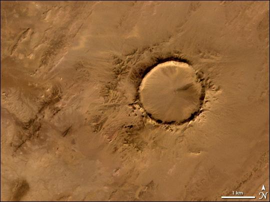 Deep in the Sahara lies a crater, a nearly a perfect circle that is 1.9 km (1.2 mi) wide, and sports a rim 100 m (330 ft) high. The Tenoumer crater sits in a vast plain of rocks so ancient they were deposited hundreds of millions of years before the first dinosaurs walked the Earth. Close examination of the structure has revealed that the crater's hardened "lava" was actually rock that had melted from a meteorite impact.
On this satellite image the crater's outline is unmistakable, yet it does not necessarily look like a crater; the light and shadows make it look more as if someone pressed a giant cookie cutter into the rock. In this image, the sunlight shines from the southeast (lower right), and the bright arc along the northwestern part of the crater is where the crater walls slope up to the rim. Around the perimeter, the relatively steep walls cast dark shadows. Although it resides in ancient rock, Tenoumer is geologically young, ranging in age between roughly 10,000 and 30,000 years old. Photo courtesy of NASA.