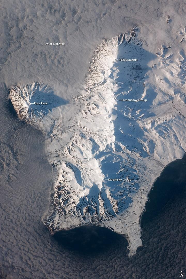 The Kuril Island chain is built from a line of volcanoes, an island arc, that extends from Russia's Kamchatka Peninsula to northern Japan. Island arcs form along an active boundary between two tectonic plates, with one being driven beneath the other (subduction). Magma generated by subduction feeds volcanoes - and eventually volcanic islands - over the subduction boundary. Paramushir Island in the northern Kurils is an example of a large island built by several volcanoes over geologic time. This astronaut photograph shows the southern end of Paramushir Island after a snowfall. The western slopes of the mountains are brightly illuminated, while the eastern slopes are in shadow.
Four major volcanic centers create this part of the island. Fuss Peak (image center left) is an isolated stratovolcano connected to the main island via an isthmus. Fuss Peak last erupted in 1854.
The southern tip of the island is occupied by the Karpinsky Group of three volcanic centers. A minor eruption of ash following an earthquake occurred on this part of the island in 1952.
The Lomonosov Group to the northeast (image center) includes four cinder cones and a lava dome.
The most recent volcanic activity on Paramushir Island occurred in 2008 at the Chikurachki cone located along the northern coastline of the island at image top center. The summit of this volcano, 1,816 m (5,958 ft) above sea level, is the highest on Paramushir Island.
Much of the Sea of Okhotsk visible in the image is covered with low clouds that often form around the islands in the Kuril chain. The clouds are generated by moisture-laden air passing over the cool sea/ocean water, and they typically wrap around the volcanic islands. Image courtesy of NASA.