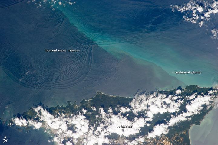 This space station photo of 18 January 2013, shows the north coast of Trinidad and a series of subtle, interacting arcs in the southeastern Caribbean Sea. These are known as "internal waves," the surface manifestation of slow waves that move tens of meters beneath the sea surface. Internal waves produce enough of an effect on the sea surface to be seen from space, but only where they are enhanced due to reflection of sunlight, or sunglint, back towards the space station.
The image shows at least three sets of internal waves interacting. The most prominent set (image top left) shows a packet of several waves moving from the northwest due to the tidal flow towards the north coast of Trinidad. Two less prominent, younger sets can be seen further out to sea. A very broad set enters the view from the north and northeast, and interacts at image top center with the first set. All the internal waves are probably caused by the shelf break near Tobago (outside the image to top right). The shelf break is the step between shallow seas (around continents and islands) and the deep ocean. It is the line at which tides usually start to generate internal waves.
Over the island of Trinidad, the heating of the land surface sets off the growth of cumulus clouds. Off the coast, a light blue northwest-southeast trending plume at image center is sediment embedded in the Equatorial Current (also known as the Guyana Current). The current is transporting material to the northwest - in almost the opposite direction of the internal waves. The current flows strongly from east to west around Trinidad, all the way from equatorial Africa, driven by year-round easterly winds. Seafarers in the vicinity of Trinidad are warned that the current - and its local reverse eddies - make navigation complicated and sometimes dangerous for smaller craft in these waters.
Astronauts also have observed internal waves in other parts of the world, such as San Francisco and the Straits of Gibraltar. Photo courtesy of NASA.