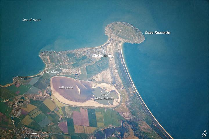 Cape Kazantyp is a prominent headland on the Kerch Peninsula, which defines the southern shore of the Sea of Azov and the east extension of the Crimean Peninsula. Due to its relatively low latitude (45° N) the Crimea has been the warm holidaying destination for generations of Ukrainians and Russians. Towns in the Kazantyp area-Lenine, Shcholkine-offer tourist attractions ranging from birdwatching to beaches and music festivals.
This detailed astronaut photograph was taken from the International Space Station when it was located 400 km (250 mi) to the northeast, but due to the long (800 mm) lens used, the photo includes detailed field patterns and city blocks. Green and brown fields show intensive agricultural activity in the area, and salt ponds are visible at the west end of the shallow Lake Aktashskoye at image center. The distance from the tip of the Cape to the largest local city, Lenine is only 20 km (12 mi).
Shcholkine is a new town built in 1978 to house workers of the planned Crimean nuclear power plant. The partially completed plant was inspected following the Chernobyl disaster of 1986 and found to be located on a site prone to earthquakes. Further construction was terminated, and the construction equipment and already built structures were abandoned in place. Today, Shcholkine is an increasingly popular tourist destination. Image courtesy of NASA.