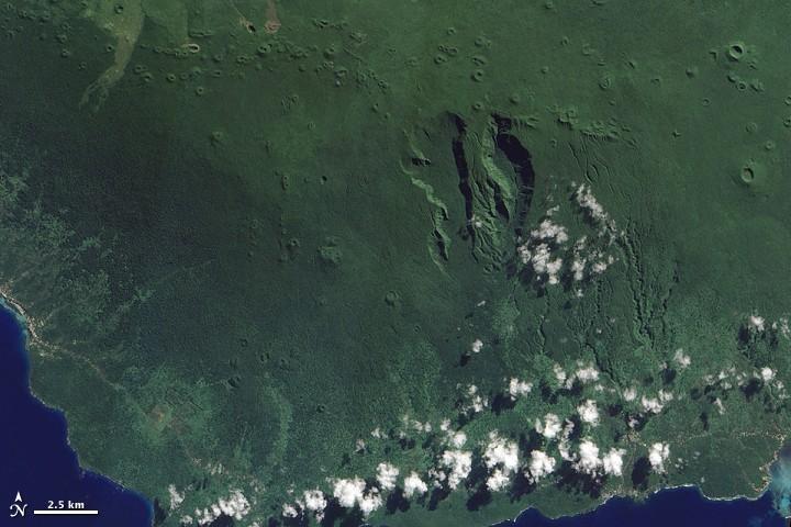 At the western end of the Samoan Island chain lies Savai'i. Stretching over 1,682 sq km (649 sq mi), Savai'i is one of the largest landmasses in Polynesia. The mountainous island is a shield volcano that reaches its highest altitude, 1,858 m (6,095 ft), near the center of the island. The region's tropical, humid climate sustains vegetation that carpets much of the island.
A jagged line of small, round volcanic craters extends east-west across the island (top of image). South of this uneven line, two deep, parallel valleys extend toward the south. The semicircular contours of the valleys suggest that they might be the remains of an ancient caldera or collapsed magma chamber that was modified by the later eruption of additional lava.
With the exception of a few brown crater rims, and a coastal settlement along the image's left edge, the landscape appears in varying shades of green. Dense rainforests cover the island's rugged interior, and thick vegetation also thrives along the coastal plains.
Savai'i has remained volcanically active into historical times, with two eruptions occurring in the early 20th century. The eruptions sent big lava flows to the island's northern coast, destroying several villages. Photo courtesy of NASA.