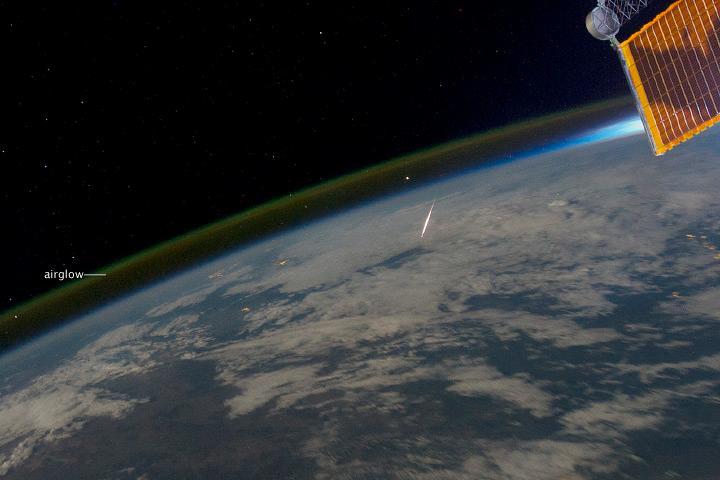 Many people have spent time outdoors under a dark sky, watching for "shooting stars" to streak across the firmament. While not actual stars, "shooting stars" do come from outer space, in the form of meteoroids entering the Earth's atmosphere. Meteoroids are small objects moving through the solar system that are attracted to the Earth by its gravitational pull. These small objects - typically fragments of asteroids or comets, though they can also originate from the Moon or Mars - begin to heat and burn up as they collide with air molecules in Earth's atmosphere, creating a bright vapor trail or streak. At this point, the object is known as a meteor. If any remnant of the object survives to impact the Earth's surface, it becomes known as a meteorite. While most meteorites are natural in origin, on occasion manmade space debris can reenter the atmosphere and also become a meteor or even a meteorite!
This astronaut photograph provides the unusual perspective of looking down on a meteor as it passes through the atmosphere. The image was taken on 13 August 2011, during the Perseid Meteor Shower that occurs every August. Green and yellow airglow appears in thin layers above the limb of the Earth, extending from image left to the upper right. Atoms and molecules above 50 km (30 mi) in the atmosphere are excited by sunlight during the day, and then release this energy at night, producing primarily green light that is observable from orbit. Part of a space station solar panel is visible at image upper right; behind the panel, a bright region indicates the Sun low on the horizon. Image courtesy of NASA.